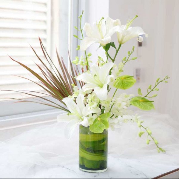 White lilies, green anthurium, and foliage in a glass vase with green accents.