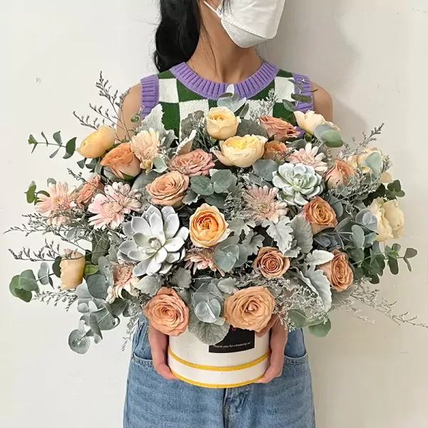 A person holds a round hat box filled with peach, pink, and cream roses, succulents, and eucalyptus.