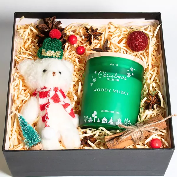 Christmas gift box with teddy bear, scented candle, pine cone, cinnamon sticks, and red ornaments.