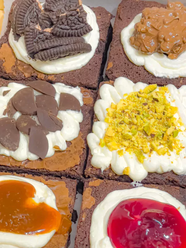 Six square brownies topped with vanilla frosting and various toppings: Oreos, chocolate, caramel, pistachios, and cherry.