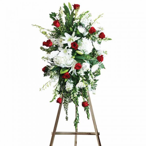Standing funeral spray of white lilies, hydrangeas, chrysanthemums, and red roses on a wooden easel.