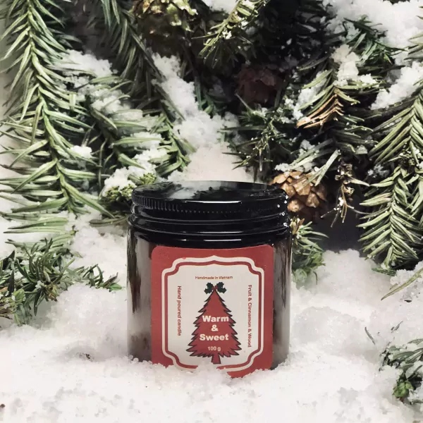 Jar candle with a red label depicting a Christmas tree and text "Warm & Sweet 100g" on snow with pine branches.