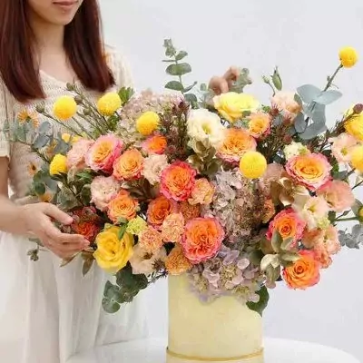 A woman arranges a large, vibrant bouquet of orange, pink, and yellow roses, hydrangeas, and yellow ping pong chrysanthemums in a pale yellow box.