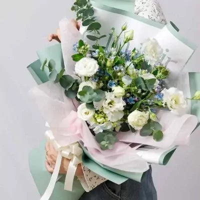 A bouquet of white lisianthus, hydrangea, and blue fillers with eucalyptus leaves, wrapped in light green and pink paper.