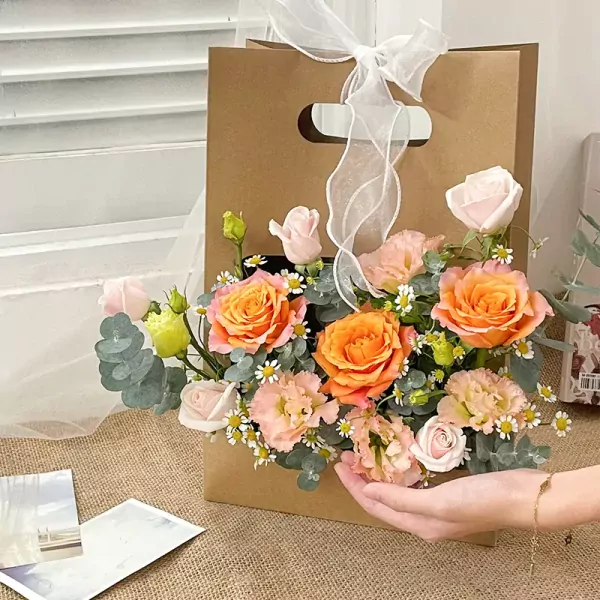Basket of orange roses, pink roses, and other flowers with eucalyptus in a brown paper bag with a white ribbon.