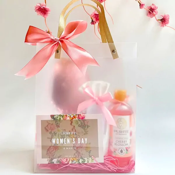 A clear gift box with pink ribbon, a floral card, a pink pouch, and a cherry blossom body wash.