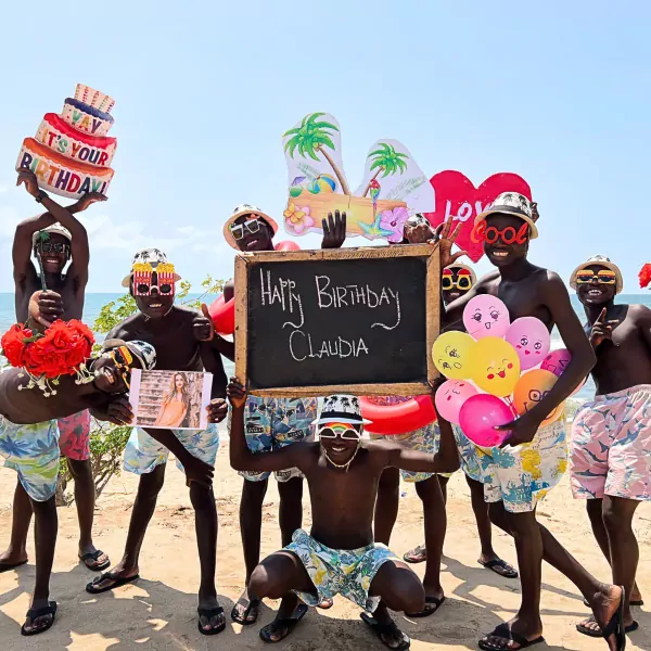 Birthday celebration on the beach with a group of smiling African men holding balloons and a "Happy Birthday Claudia" sign.