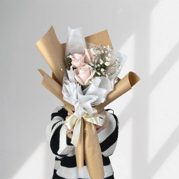 A bouquet of three light pink roses and baby's breath, wrapped in brown and white paper with a beige ribbon.