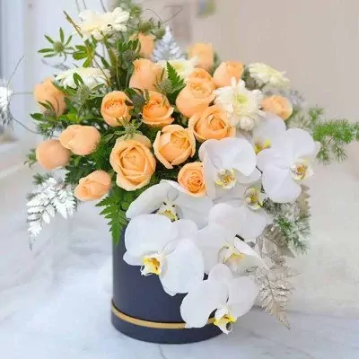 Peach roses, white orchids, and greenery in a dark round box.