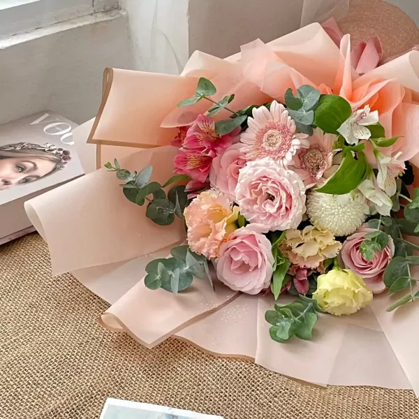A pastel pink floral bouquet featuring roses, daisies, and other delicate blooms, wrapped in light pink paper.