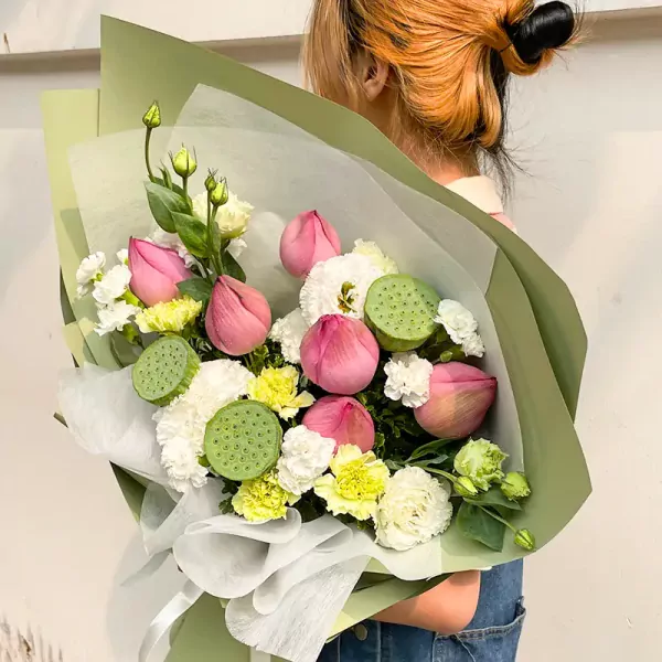 A bouquet of pink lotus flowers, green lotus seed pods, and white carnations wrapped in green paper.