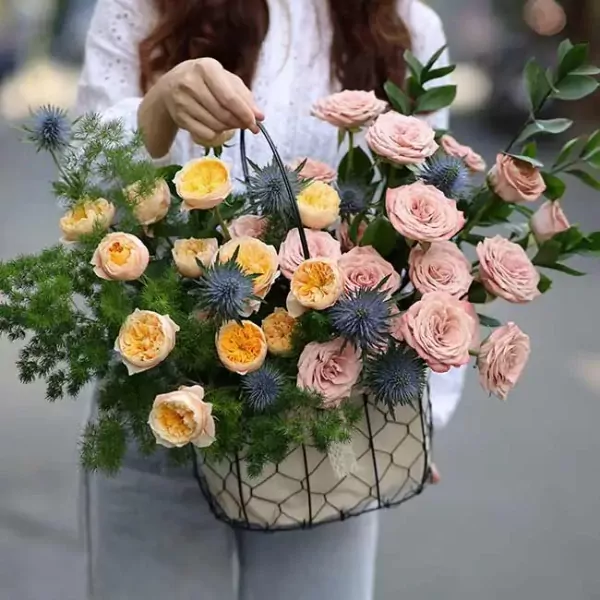 Basket of pink and yellow roses, blue thistles, and green foliage.