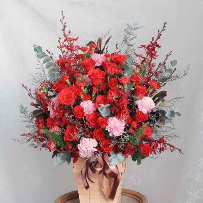 Large floral arrangement of red roses, carnations, and greenery in a woven basket with a ribbon.
