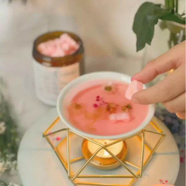 Hand adding pink wax melt cubes to a pink liquid in a white bowl over a candle warmer.