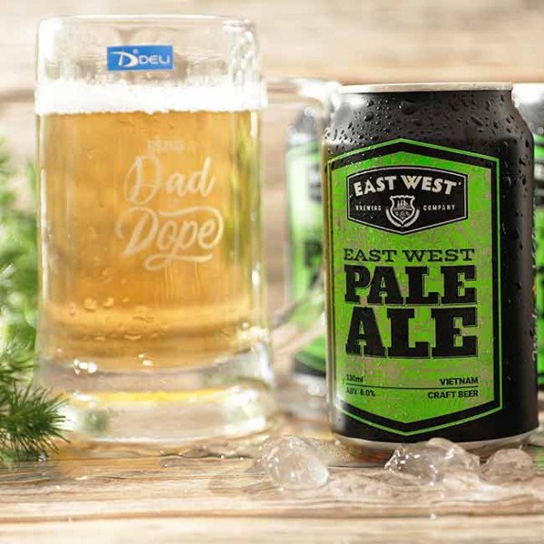 Glass mug with "Being Dad Is Dope" text next to an East West Pale Ale beer can.