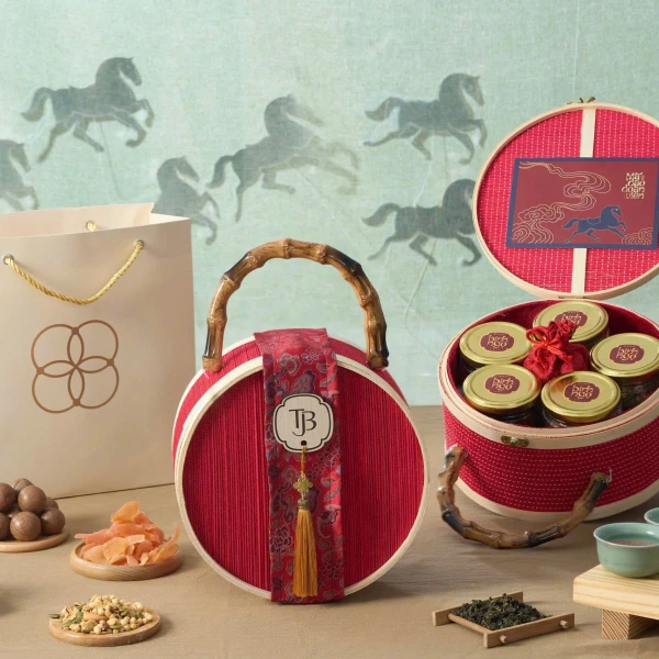 Round red gift basket with bamboo handle, containing jars of nuts, dried fruit, and tea.