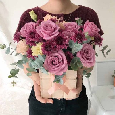 A woman holds a box of pink roses, purple chrysanthemums, and eucalyptus leaves.
