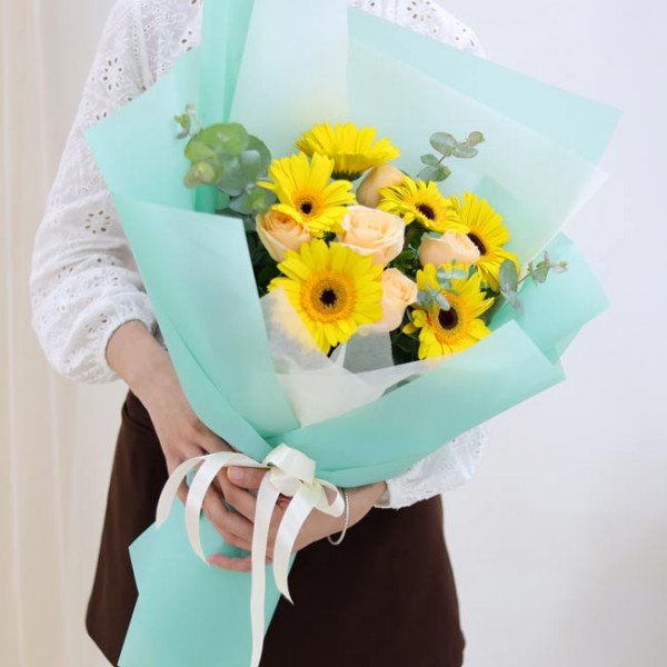 A bouquet of yellow gerbera daisies and peach roses, wrapped in aqua paper, tied with a white ribbon.