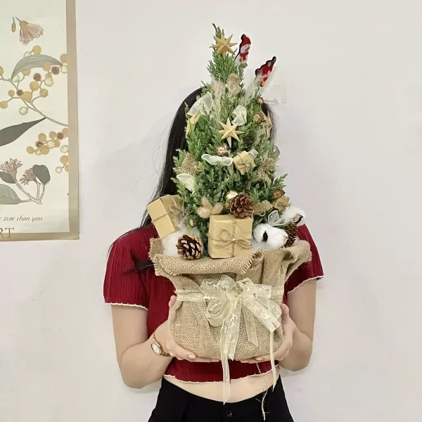 Mini Christmas pine tree decorated with pinecones, gifts, and bows in a burlap bag.