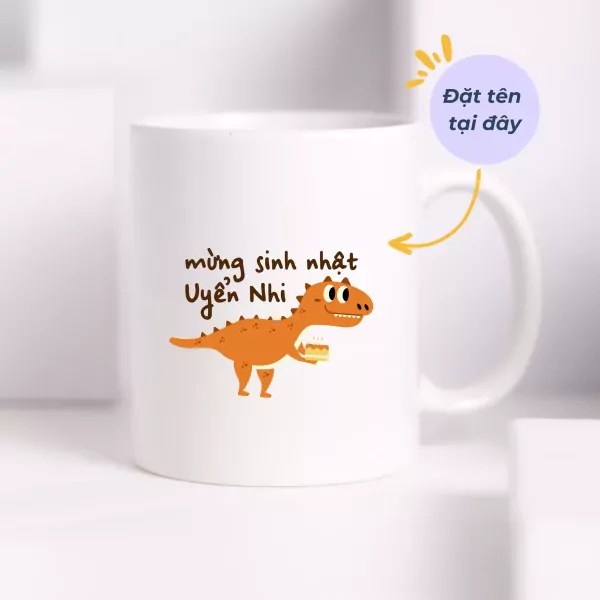 White mug with a cartoon dinosaur holding a cake and "Happy Birthday Uyen Nhi" text.