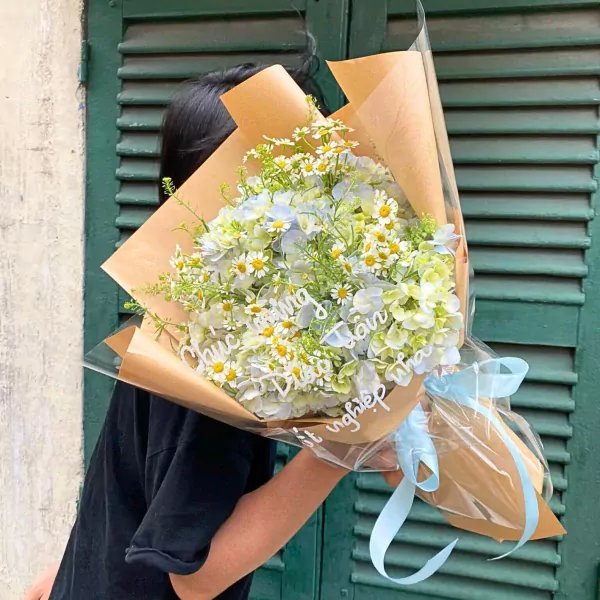 Bouquet of white and light blue flowers with daisies and greenery, wrapped in brown paper and clear plastic with a blue ribbon.