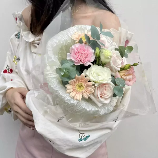 A woman holding a pastel pink and white flower bouquet, wrapped in sheer white paper with a ruffled edge.