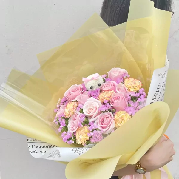 Pink roses and carnations arranged in a bouquet with a teddy bear, wrapped in yellow paper.