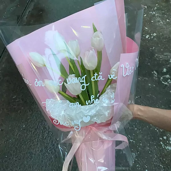 A bouquet of white tulips wrapped in pink paper and clear plastic, with Vietnamese text.