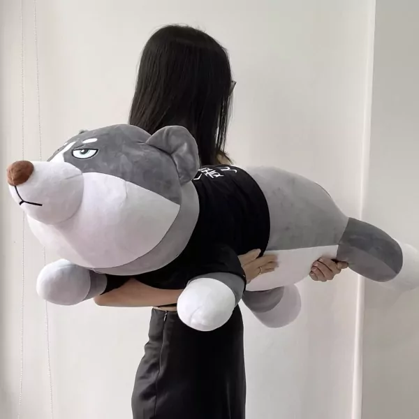 A person holds a large, grey plush husky with a grumpy expression, wearing a black shirt.