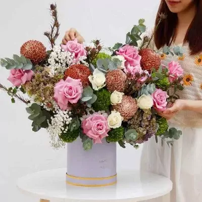 A large floral arrangement in a pastel purple box with pink roses, brown and green chrysanthemums, and other greenery.
