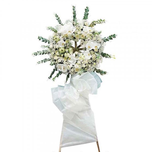 White floral wreath with orchids, roses, and greenery on a stand with a white bow.
