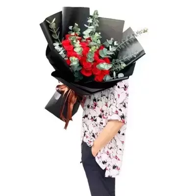 A bouquet of red roses and eucalyptus wrapped in black paper, held by a person.