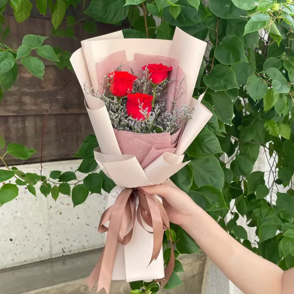 Hand holding a bouquet of 3 red roses with delicate fillers, wrapped in light pink and cream paper.