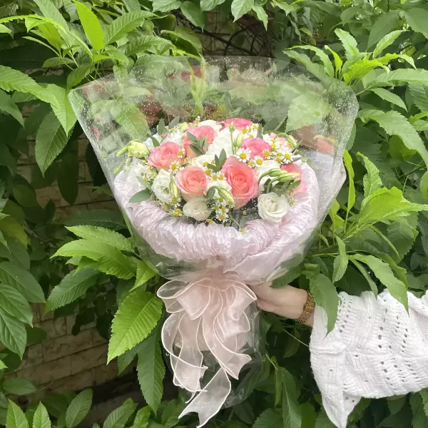 Pink and white roses and daisies in a bouquet wrapped in cellophane and pink fabric with a ribbon.