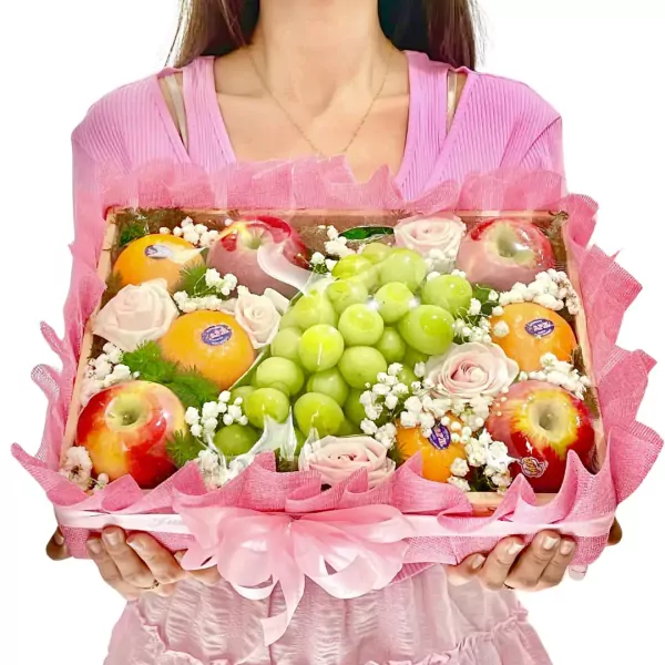 Fruit basket with green grapes, oranges, apples, and pink roses, presented by a woman in a pink top.