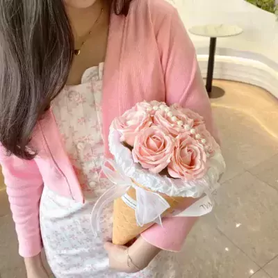 Woman in pink cardigan and floral dress holds a bouquet of pink roses in a cone.
