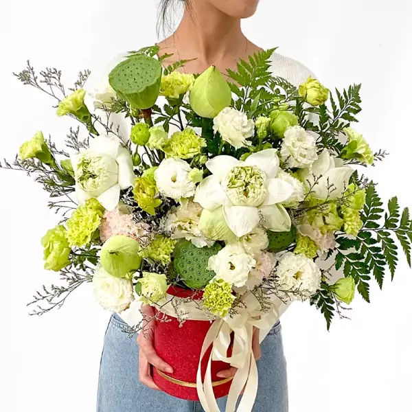 A person holds a red box filled with white lotus, green carnations, lisianthus, and lotus seed pods.