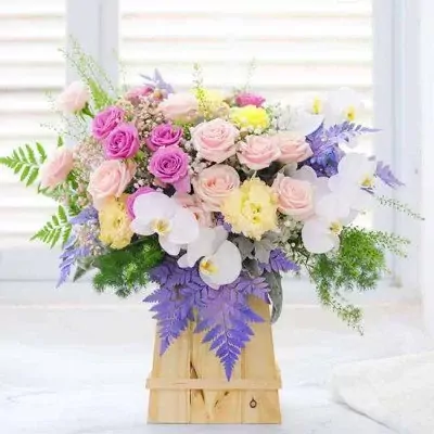 A wooden box filled with pink, purple, and yellow roses, white orchids, and green foliage.