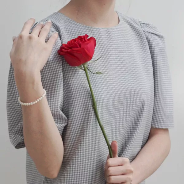 Woman in grey gingham dress wearing a pearl bracelet holds a red rose.