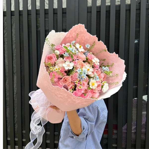 A bouquet of pink roses, carnations, blue star flowers, and white daisy-like flowers in pink paper.