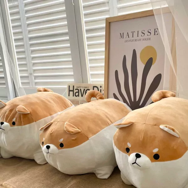 Three chonky Shiba Inu plush toys with tan and white fur, arranged on a textured surface in front of a window and art print.