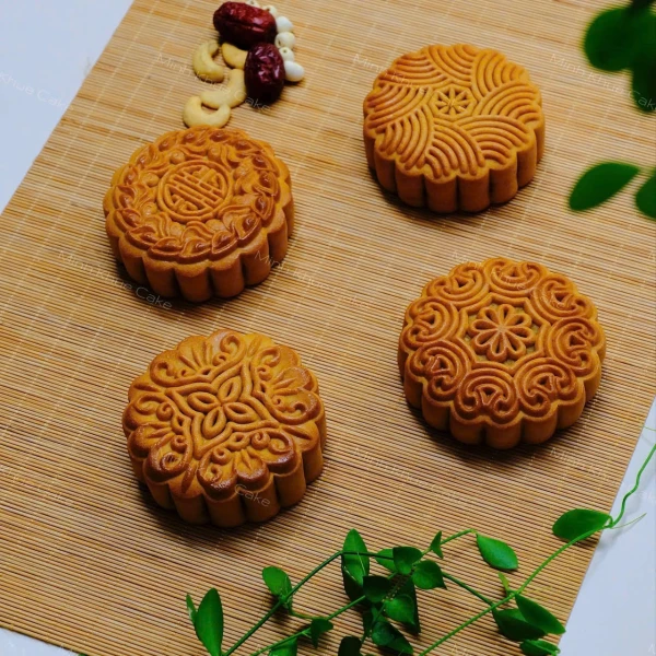 Four golden mooncakes with intricate patterns arranged on a bamboo mat, with dried fruits and nuts in the background.