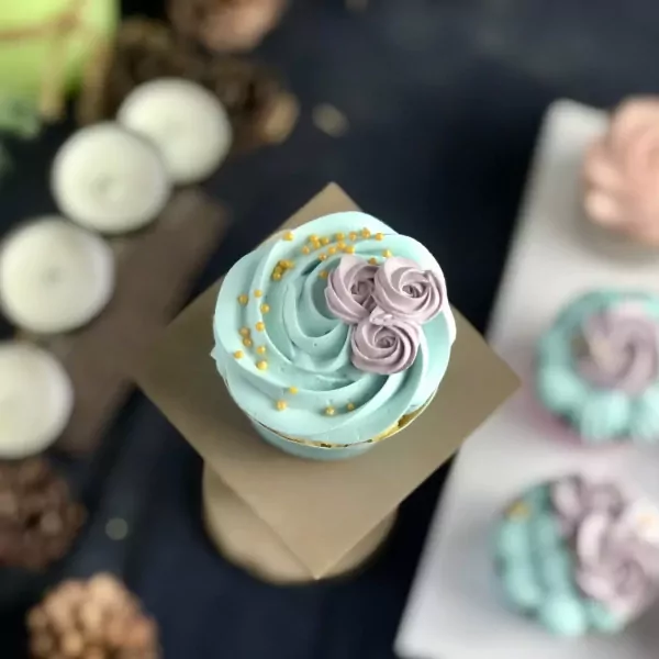 Cupcake with teal frosting, purple roses, and gold sprinkles on a stand.