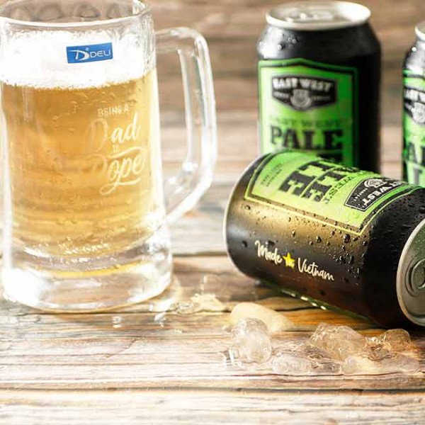Glass beer mug with "Being Dad Is Dope" text next to beer cans.