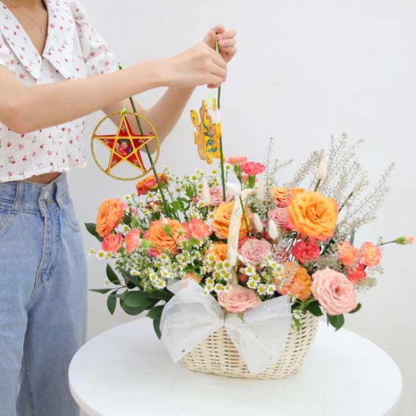 Hand holding lanterns over a flower basket with orange and pink roses, carnations, and daisies.