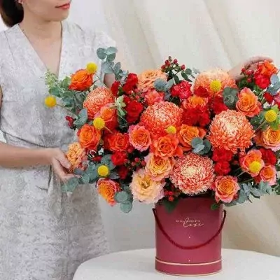 A woman holds a round, maroon hat box overflowing with bright orange roses, chrysanthemums, and yellow billy balls.