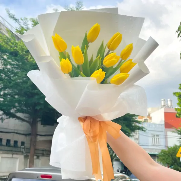 A bouquet of 10 vibrant yellow tulips, wrapped in white paper and tied with an orange ribbon.
