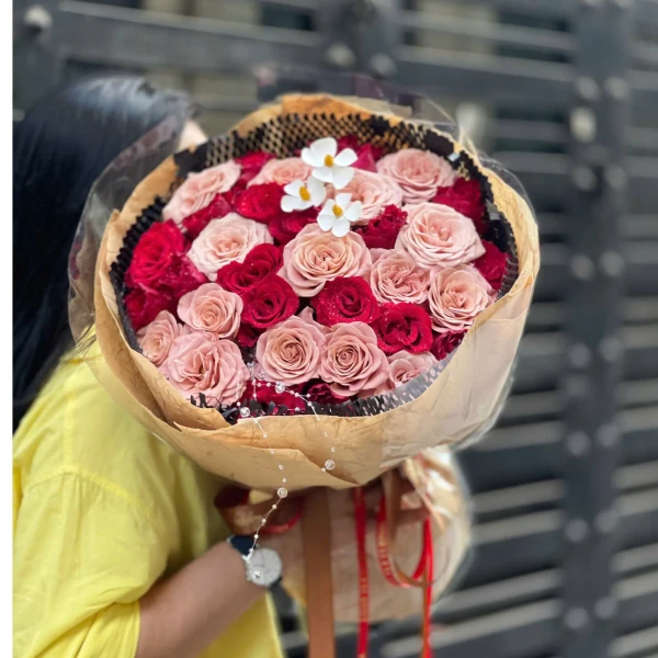 A bouquet of pink and red roses with small white flowers and decorative beads, held by a person in a yellow shirt.