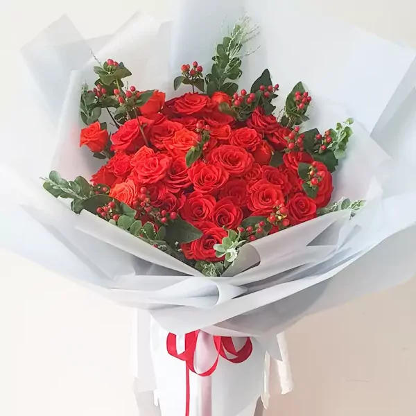 A large bouquet of red roses with green foliage and red berries, wrapped in white paper and tied with red ribbon.