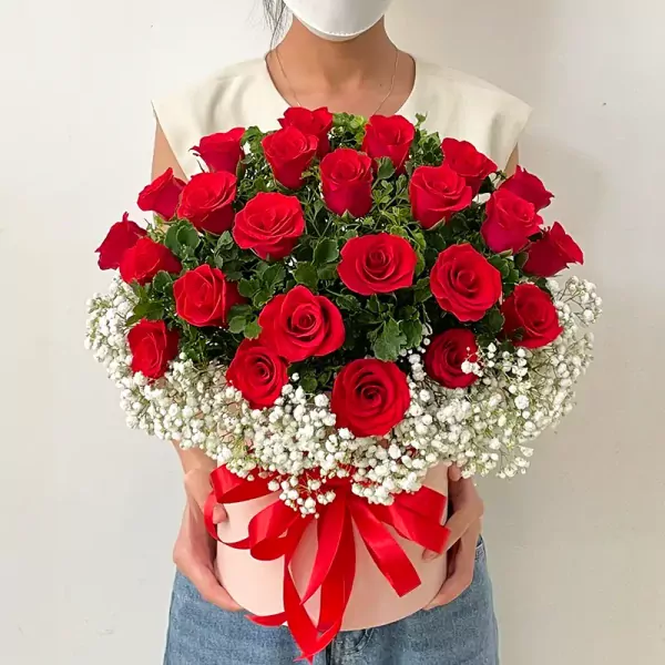 A round pink box filled with red roses and baby's breath, tied with a red ribbon.